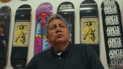 Douglas Miles Sr. standing in front of a wall of skateboards he designed for Apache Skateboards.