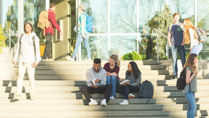 An image of college students sitting on steps in front of a campus building with other students walking nearby