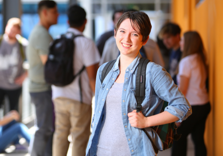 An image of a female college smiling with a backpack on one shoulder with other students in the background.