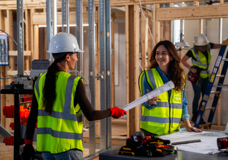Kate Glantz, Move Over Bob co-founder and CEO, is wearing a safety vest while working on a construction site with another woman construction worker who is wearing a hardhat and safety vest and is holding blueprints.
