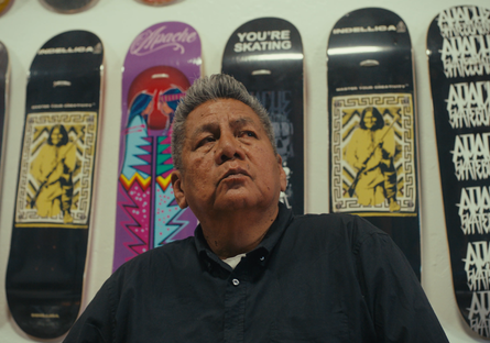 Douglas Miles Sr. standing in front of a wall of skateboards he designed for Apache Skateboards.