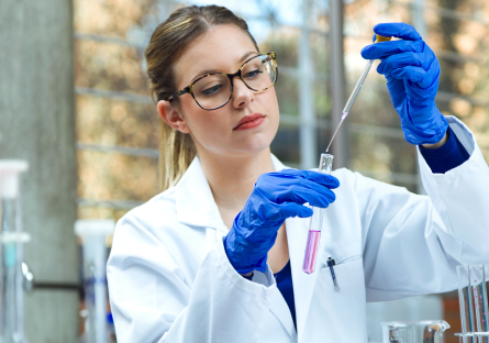 An image of a woman working in a lab wearing glasses, white lab coat, and gloves holding a beaker and dropper 