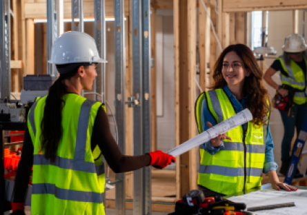 Kate Glantz, Move Over Bob co-founder and CEO, is wearing a safety vest while working on a construction site with another woman construction worker who is wearing a hardhat and safety vest and is holding blueprints.