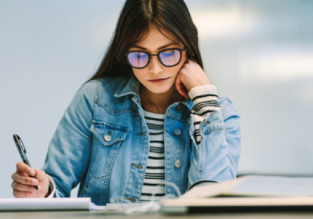 Image of a woman wearing glasses and a jean jacket holding a pen and reading a textbook and cup of coffee next to her
