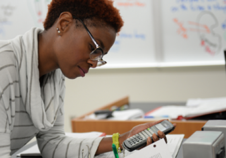 Image of woman wearing glasses and a stripped sweater using a calculator and writing in a notebook